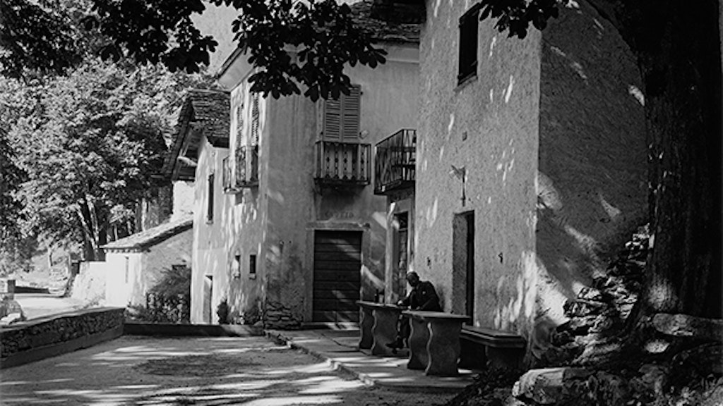 A wide shaded path leads left past a row of stone houses with grottos. In front of one house, a man sits at a stone table, barely visible. On the right side of the road stands a chestnut tree, its branch reaching across the street.