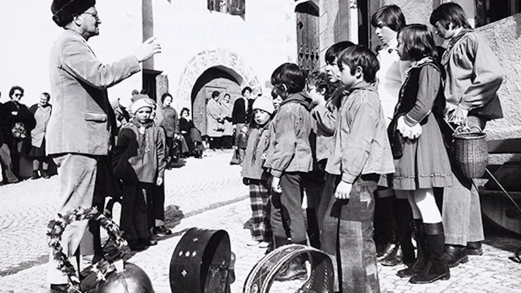 Un petit groupe d’écoliers et d’écolières en costume traditionnel accompagnés de leur enseignant s’apprêtent à chanter devant des maisons massives typiques de Guarda. Au premier plan, colliers et sonnailles indiquent que c’est le jour du Chalandamarz. À l’arrière-plan, on aperçoit le public.