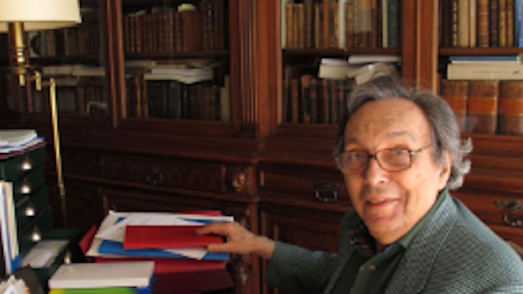 La photographie montre Jean Starobinski assis à sa table de travail, au milieu de sa bibliothèque, avec des livres ouverts devant lui.