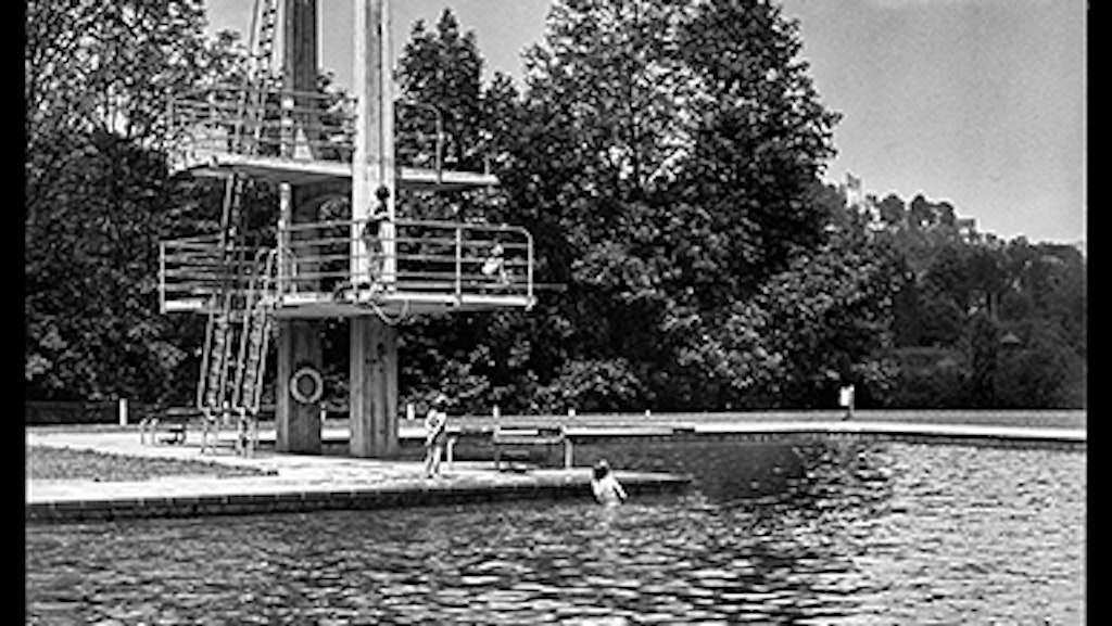 A diver performing a swan dive from the diving board of an open-air pool