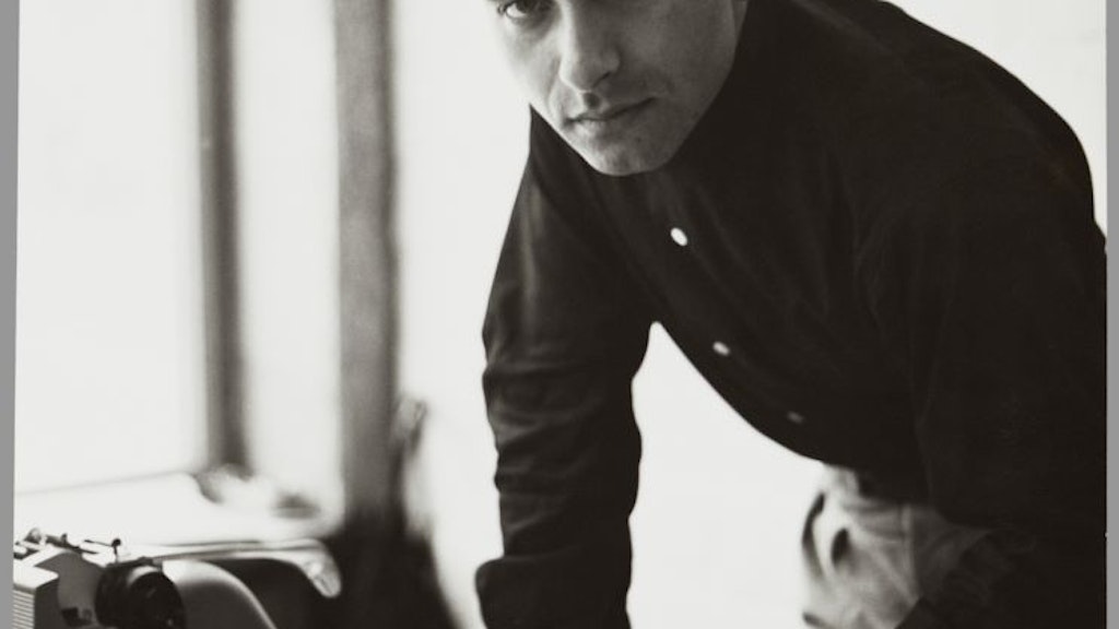 Portrait d’Eugen Gomringer, penché sur une table avec une machine à écrire, tenant un feuillet dans sa main, regardant l’appareil photo.