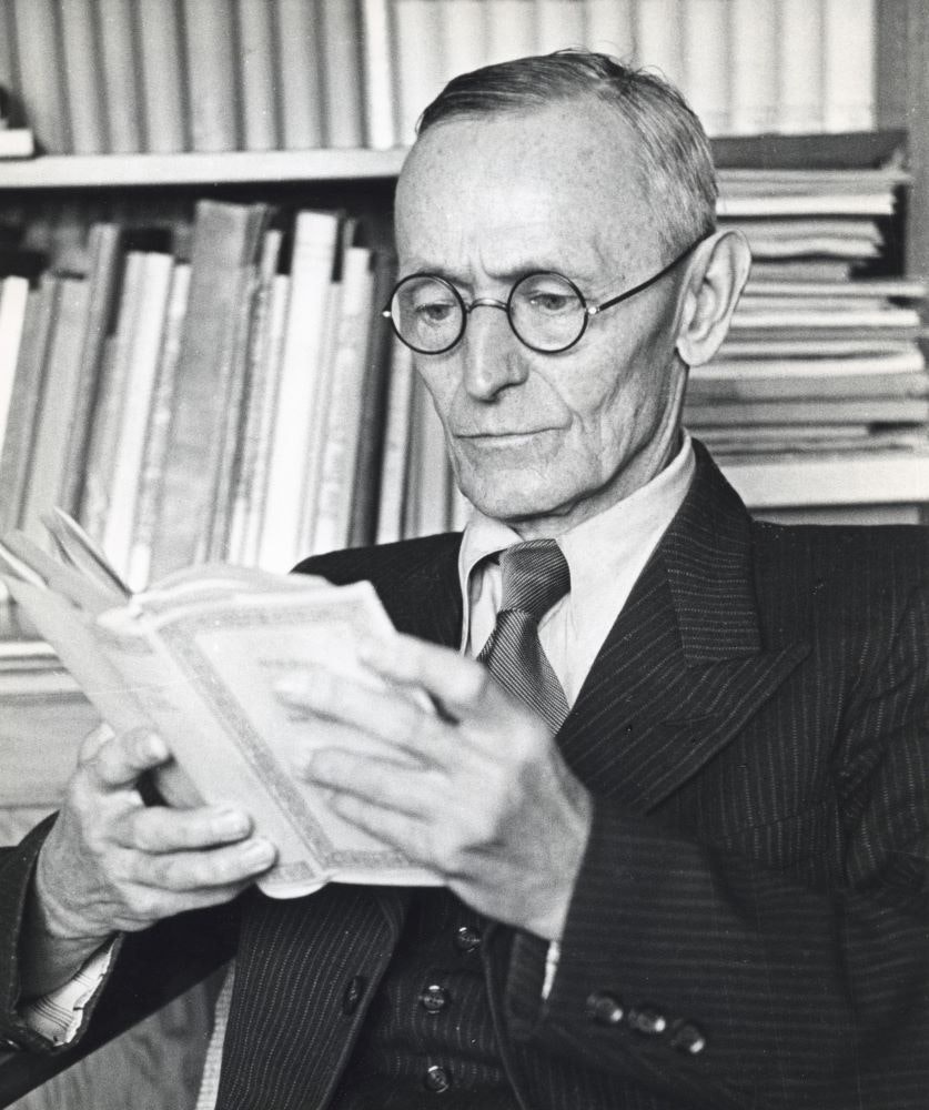 Hermann Hesse vor Bücherregal, nachdenklich in Buch vertieft.
