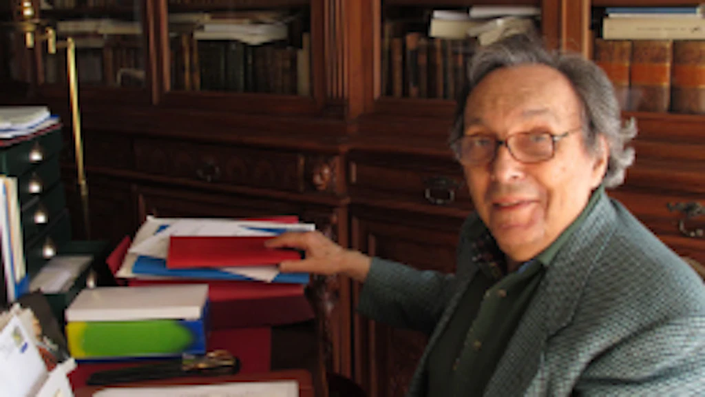 La photographie montre Jean Starobinski assis à sa table de travail, au milieu de sa bibliothèque, avec des livres ouverts devant lui.