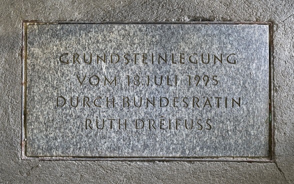 Die Inschrift auf der Bodenplatte aus grau meliertem Stein erinnert an die Grundsteinlegung vom 18. Juli 1995 durch Bundesrätin Ruth Dreifuss.