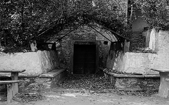 Frontansicht eines zweigeschossigen Grottos, umgeben von Bäumen, die noch ohne Laub sind. Auf Strassenhöhe die geschlossene Eingangstür zum Grotto, am Boden trockene Laubblätter. Links und rechts vom Eingang ein Steinmäuerchen, das zum Sitzen einlädt.