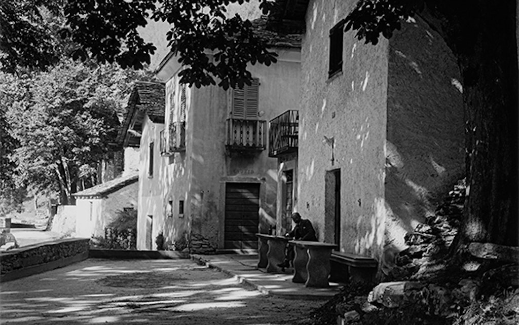 A wide shaded path leads left past a row of stone houses with grottos. In front of one house, a man sits at a stone table, barely visible. On the right side of the road stands a chestnut tree, its branch reaching across the street.