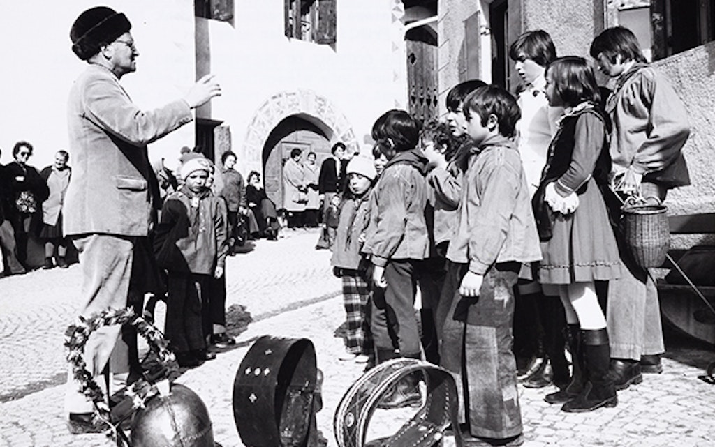A small group of schoolchildren in traditional dress are standing with their teacher in front of the typical imposing buildings in Guarda, ready for their singing performance. The bells in the foreground indicate Chalandamarz. The public can be seen in the background.