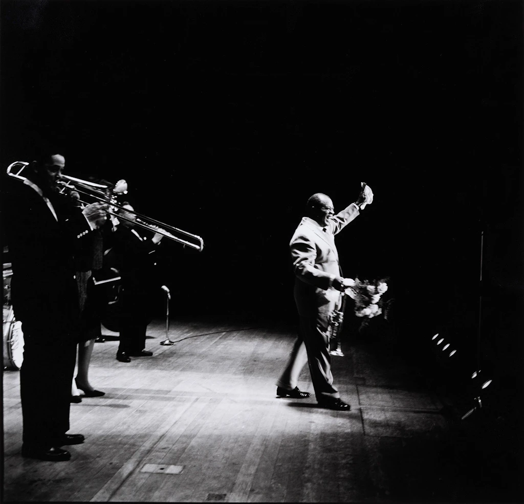 Louis Armstrong on stage holding a bouquet of flowers and greeting the audience; behind him are two members of the group