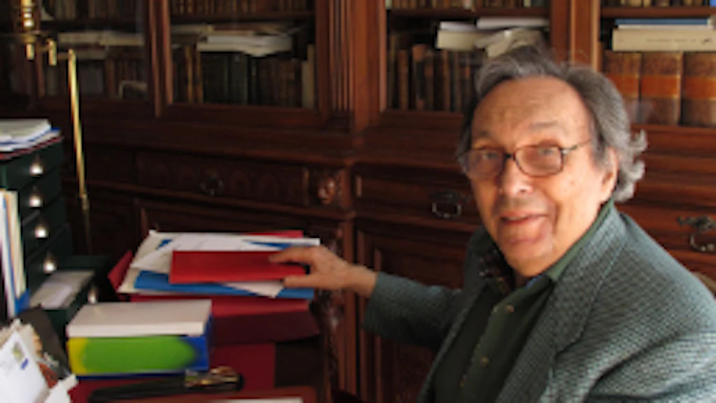 La photographie montre Jean Starobinski assis à sa table de travail, au milieu de sa bibliothèque, avec des livres ouverts devant lui.