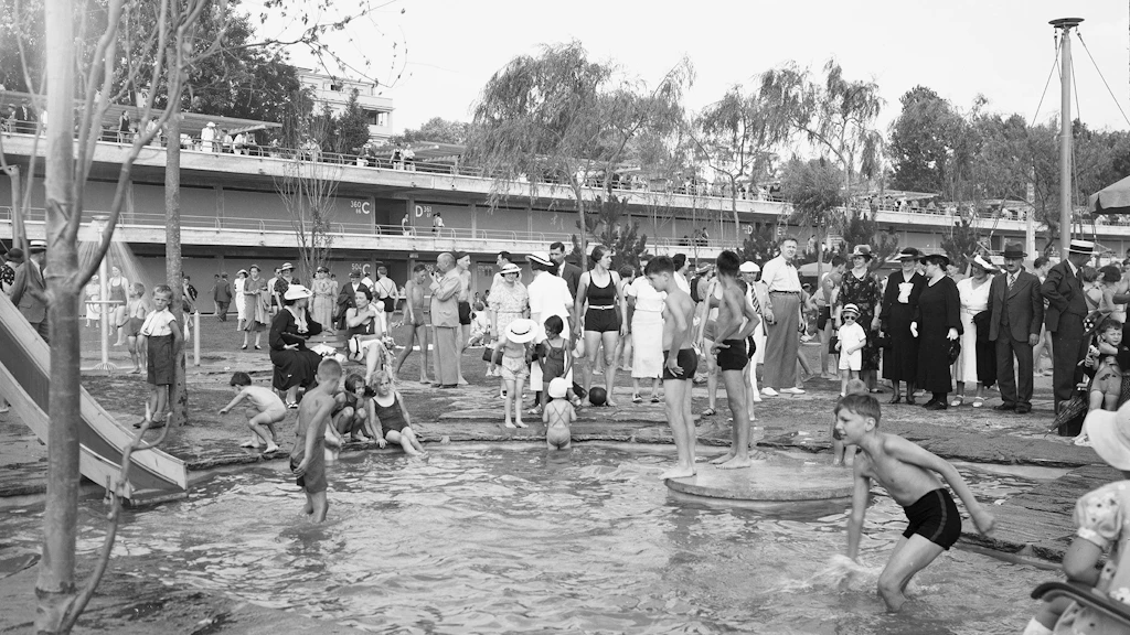 The black and white photograph shows bathers at the Bellerive Plage lido in Lausanne, circa 1940. In the foreground children splash about and enjoy themselves in a small pool, while adults of different ages stand a slight distance away. In the background we can see some of the concrete architecture of the lido, which opened in 1937. 