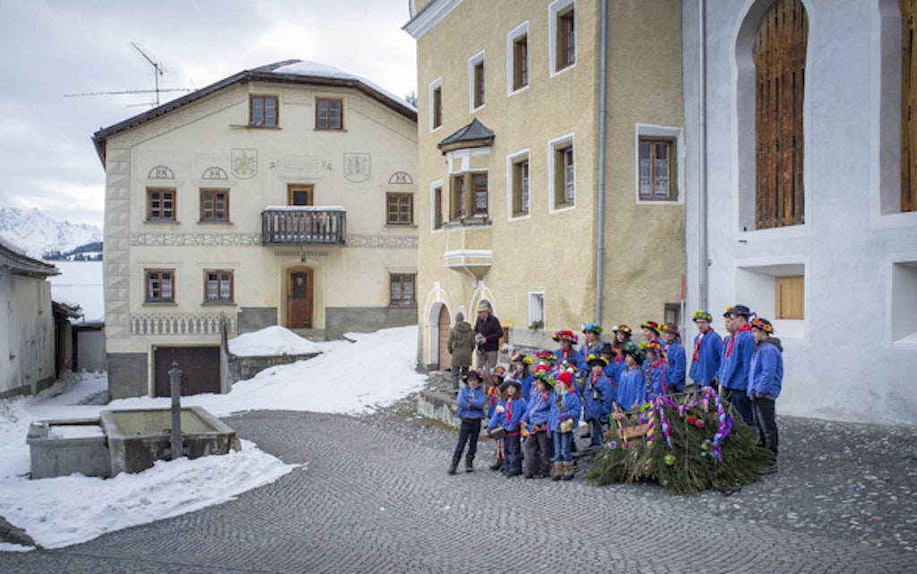 Schoolchildren sing Chalandamarz songs wearing blue smocks in front of the village fountain.