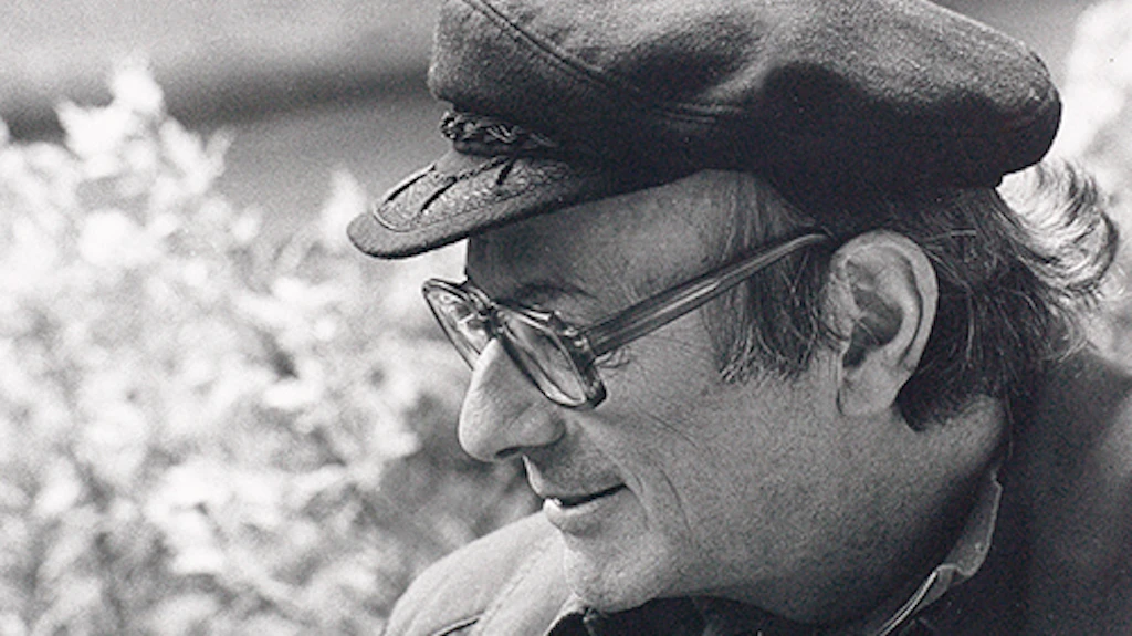 Walter Vogt wearing a peaked cap and looking downwards to the left, smiling, with a blossoming bush in the background.