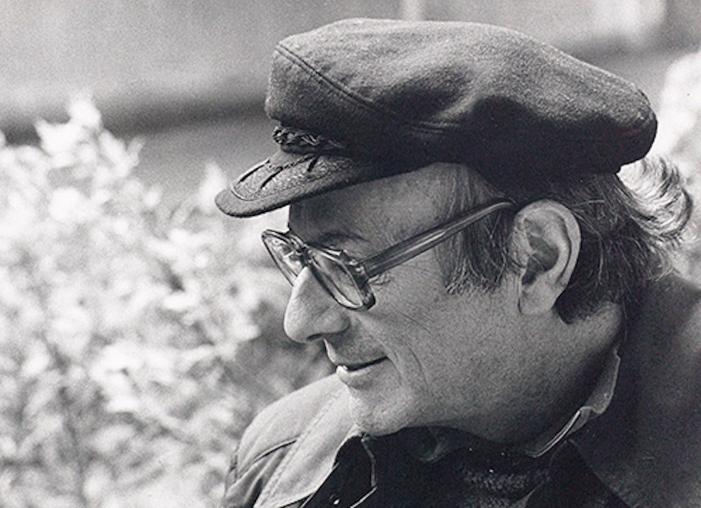 Walter Vogt wearing a peaked cap and looking downwards to the left, smiling, with a blossoming bush in the background.