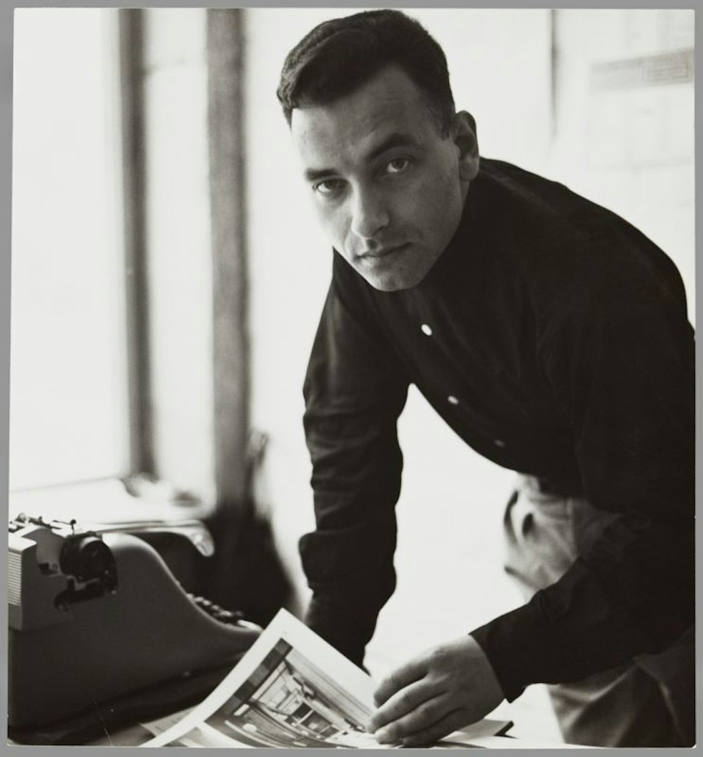 Portrait d’Eugen Gomringer, penché sur une table avec une machine à écrire, tenant un feuillet dans sa main, regardant l’appareil photo.