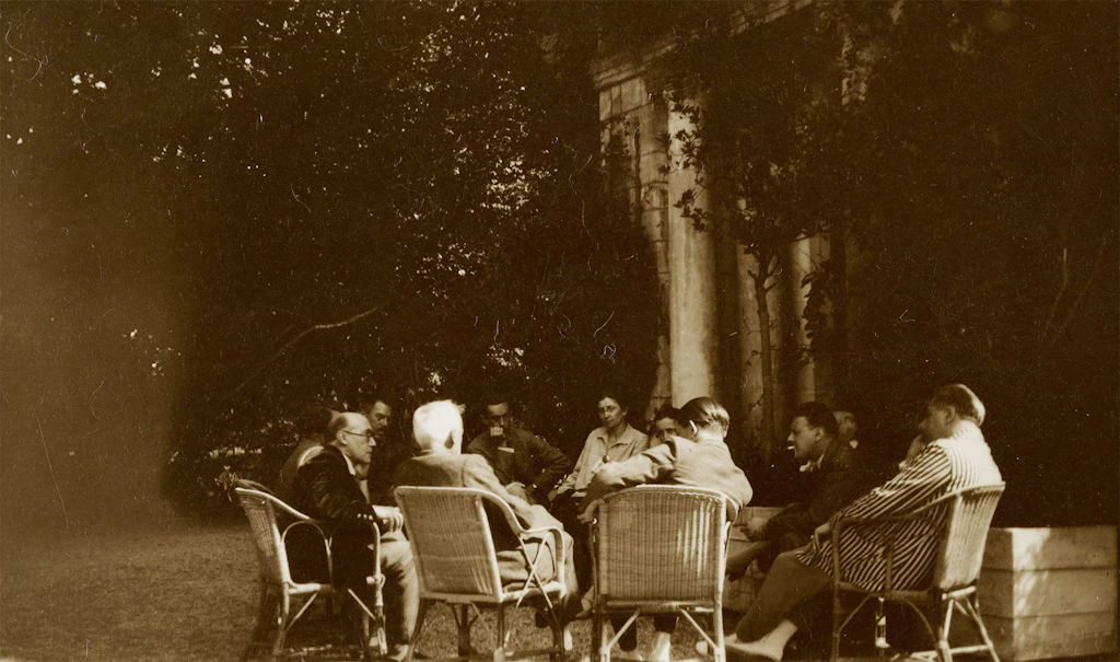 Personnes assemblées, dont André Gide, assis à gauche de profil, Léopold Chauveau, assis de dos (cheveux blancs), André Malraux, assis de dos (tourné vers Gide), Roger Martin du Gard, assis à droite, de profil (veste à rayures), et des anonymes, août 1928.