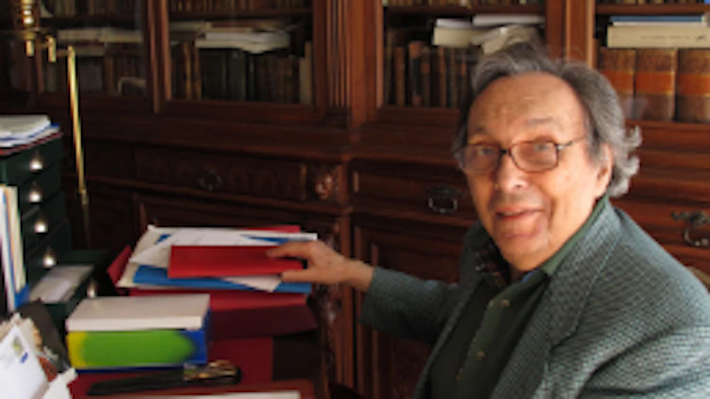 La photographie montre Jean Starobinski assis à sa table de travail, au milieu de sa bibliothèque, avec des livres ouverts devant lui.