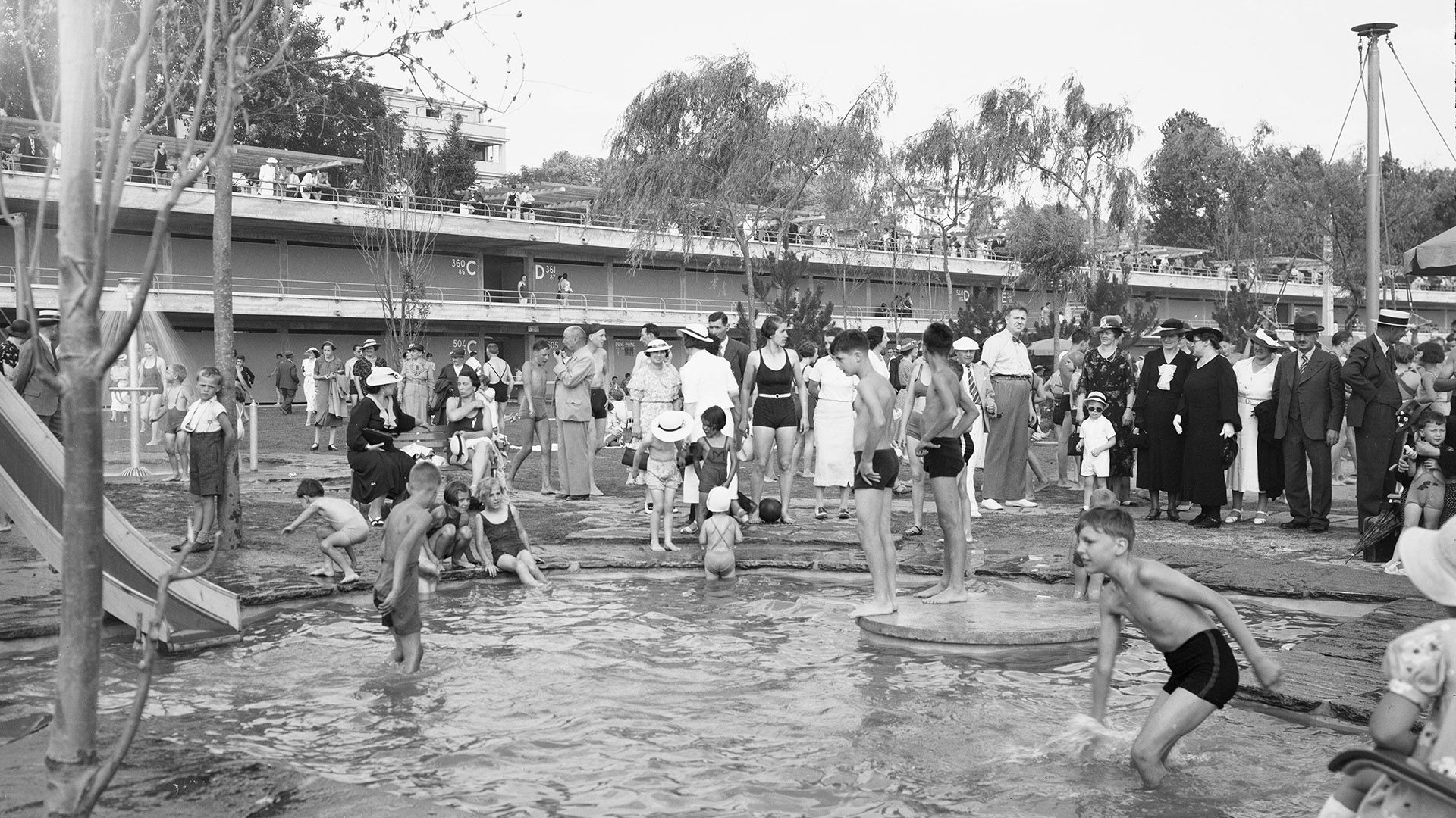 Cette photographie en noir et blanc montre des baigneurs à Bellerive-Plage, à Lausanne, vers 1940. Au premier plan, des enfants barbotent joyeusement dans un petit bassin ; un peu plus loin se tiennent des adultes de différents âges. À l’arrière-plan, on aperçoit une partie de l’architecture en béton de l’établissement, inauguré en 1937.
