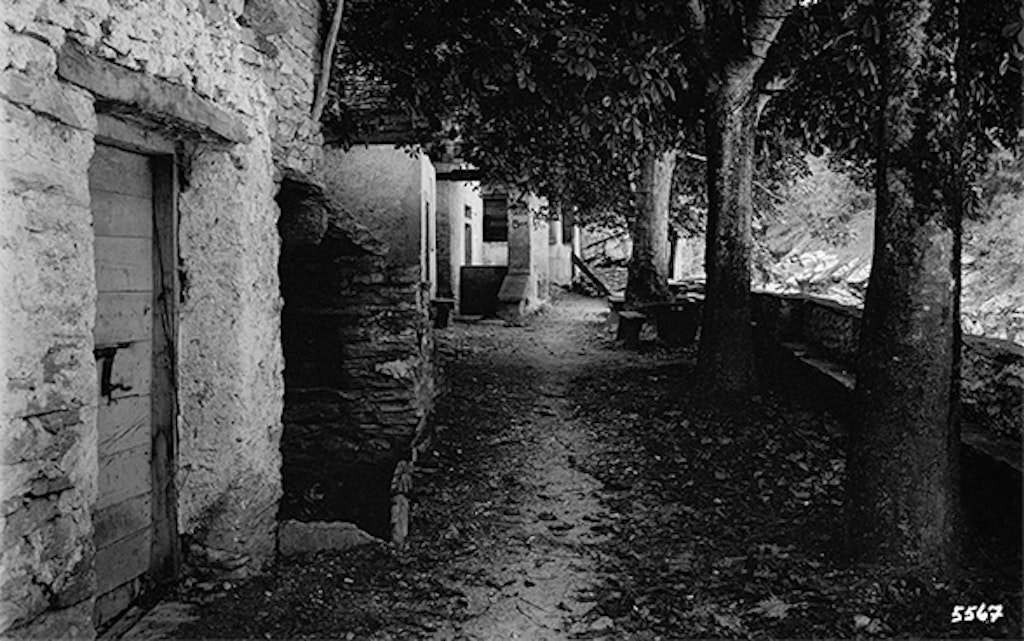 Plusieurs grotti se succèdent sur le côté gauche d’un chemin ombragé. On distingue les entrées de ces bâtiments de pierre ainsi qu’un escalier en direction de la pente. Sur la droite du chemin, on voit des châtaigniers et des feuilles mortes au sol, derrière se dresse un mur de pierre.