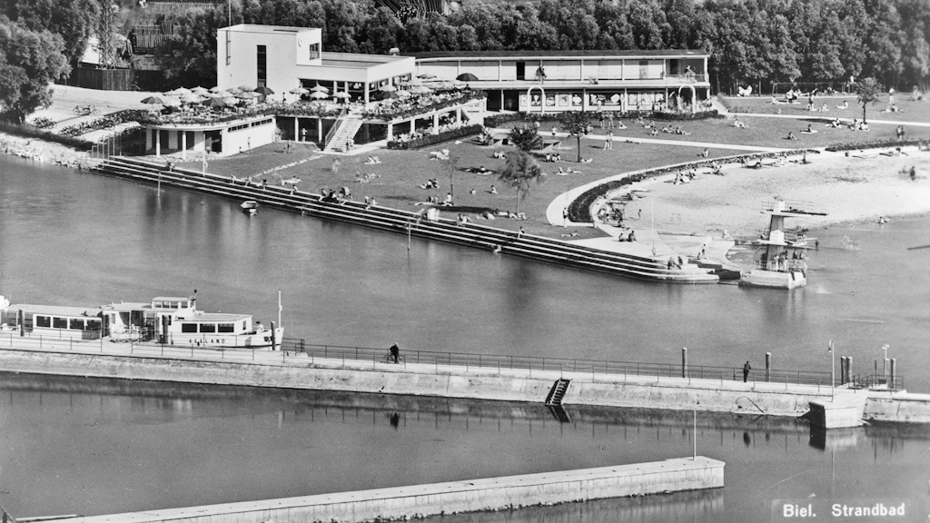 Cette photographie aérienne montre l’établissement de baignade de Bienne, construit dans le style architectural moderne du Neues Bauen des années 1930. Les bâtiments du restaurant et de l’aile des vestiaires sont dissociés l’un de l’autre. Devant eux s’étendent la pelouse et la rive du lac proprement dite. Au premier plan, on voit le bateau à moteur «Seeland» à quai.