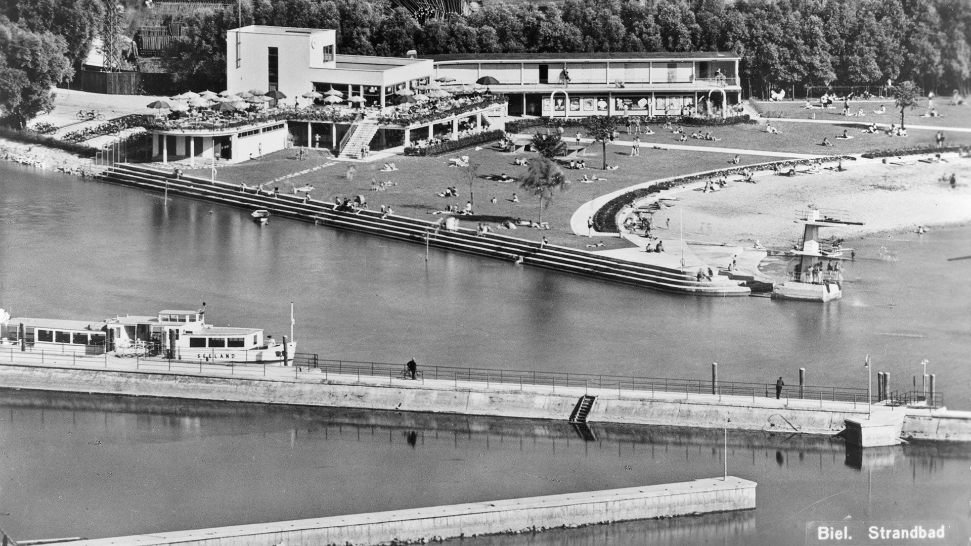 Questa veduta aerea immortala l’edificio dello stabilimento balneare di Bienne. Il complesso è composto da un fabbricato principale con il ristorante e da un’ala separata con gli spogliatoi, entrambi costruiti nello stile del Movimento moderno. Nell’area antistante si trovano il prato su cui prendere il sole e la riva del lago vera e propria. In primo piano si nota la motonave «Seeland» ormeggiata.